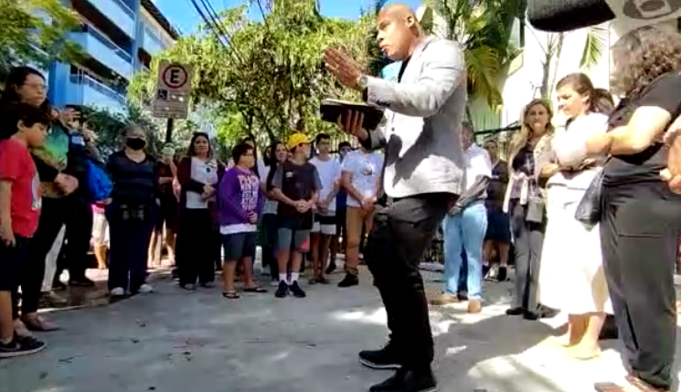 Com a Bíblia na mão, pastor fala aos participantes da reunião na porta da escola que sofreu tentativa de massacre. (Captura de tela/Tribunaonline)